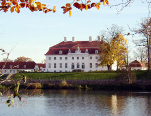 Schloss Mseberg, Ansicht von der Seeseite mit neu geweißter Fassade und rotgedecktem Dach, Ins Bild ragen mit Herbslaub bedeckte Zweige.